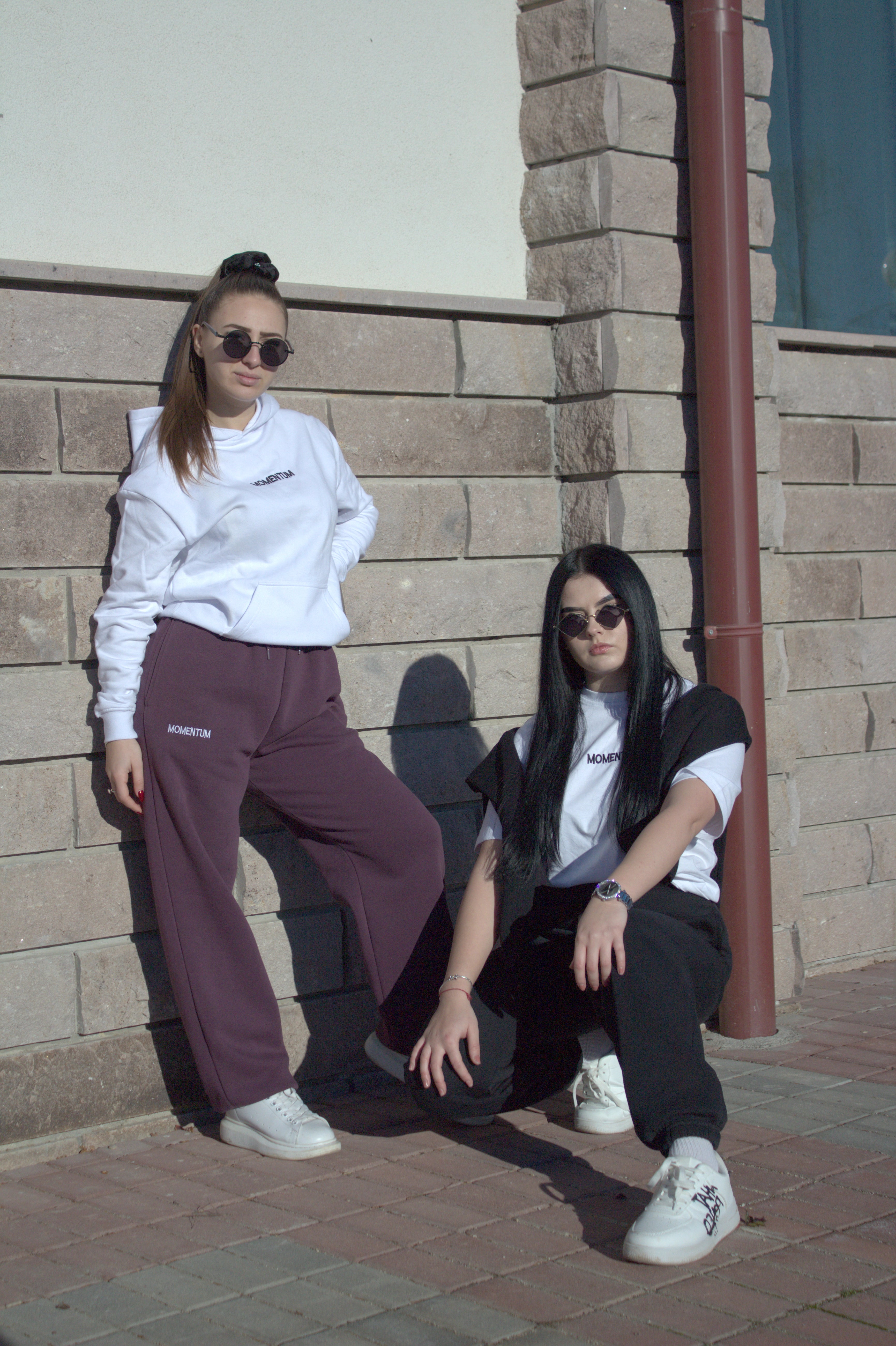 Two women posing against a brick wall, wearing sunglasses and casual clothing.